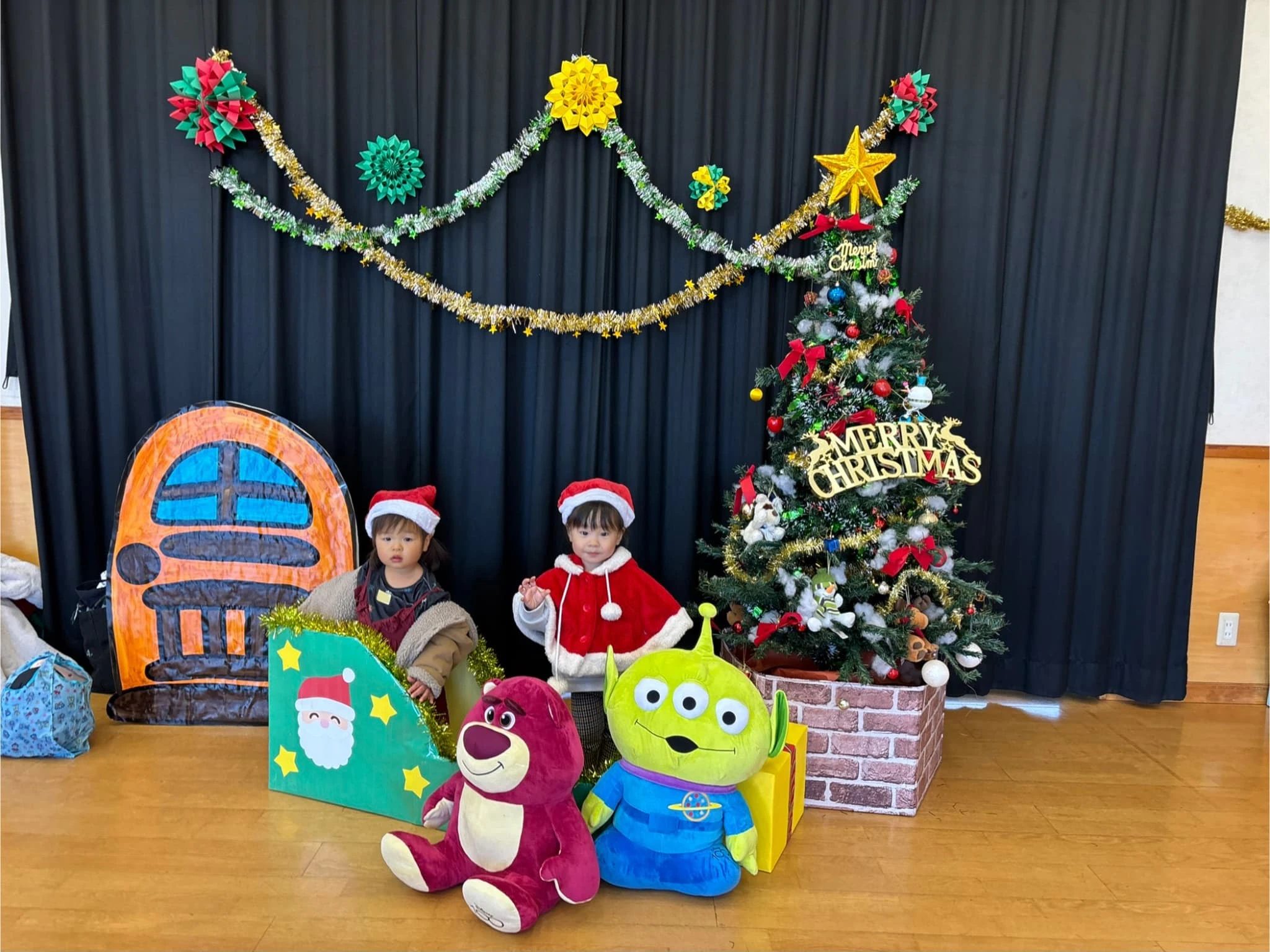 2歳の女の子二人がクリスマスツリーの前でソリに乗り写真撮影をしている様子