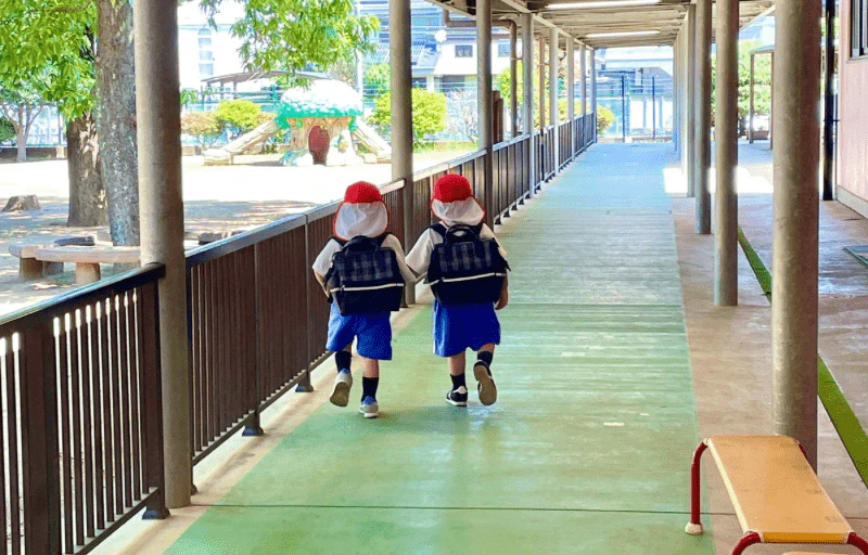 赤いカラー帽子を被った2人の年少さんが手を繋いで仲良くく後ろ姿