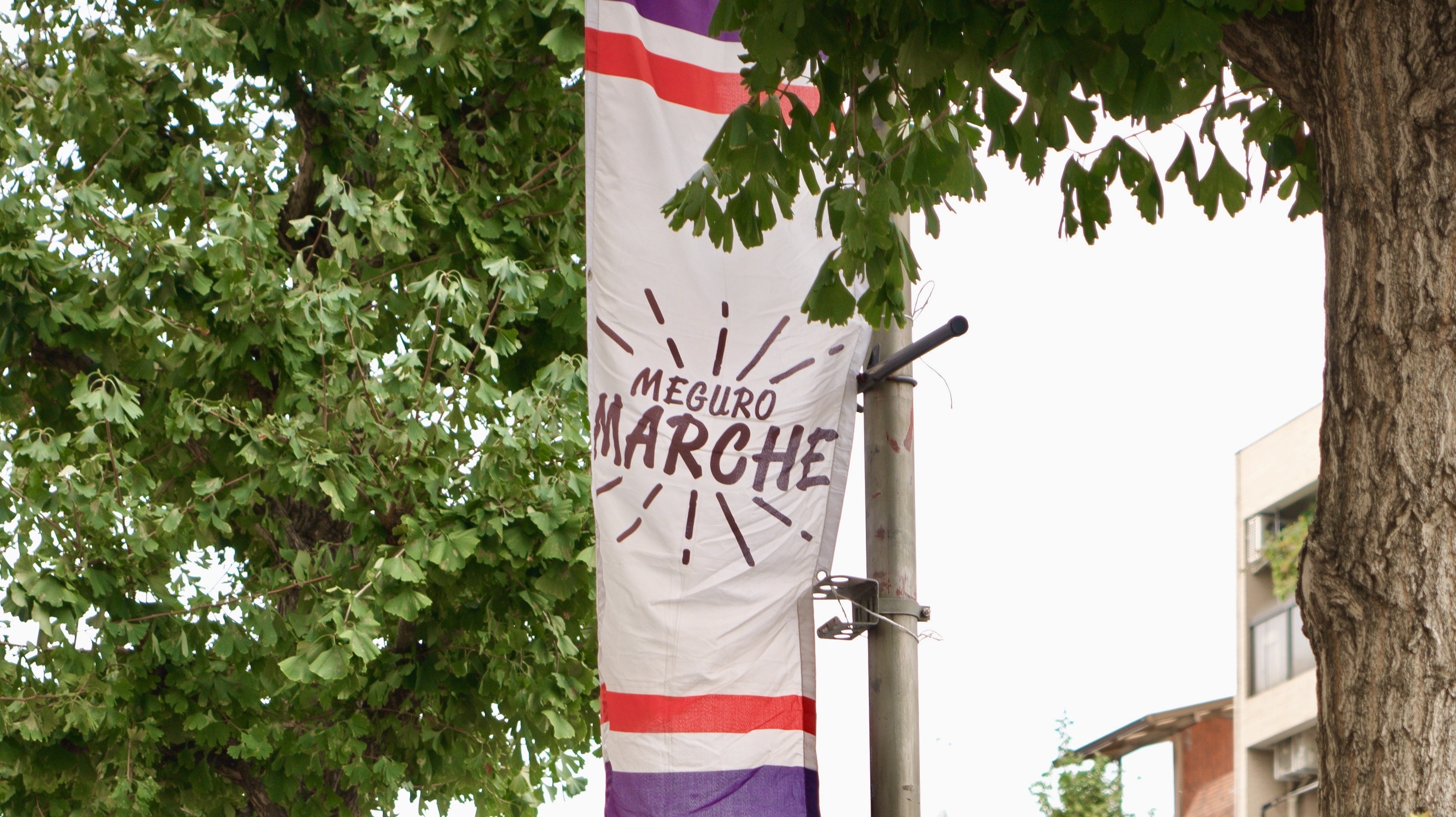太い木と緑の葉に囲まれた屋外で、白い布地に赤と紫のラインが入った縦長のバナーが掲げられている。中央には「MEGURO MARCHE」と茶色い文字で書かれている。
