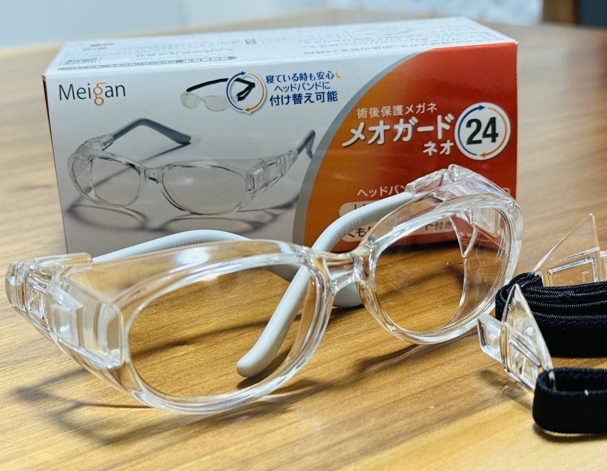 クリアなプラスチック製の保護メガネ「メオガードネオ」が木製のテーブルの上に置かれており、その奥に同じ製品のパッケージが見える。保護メガネは頭に固定するためのバンドも付属している。