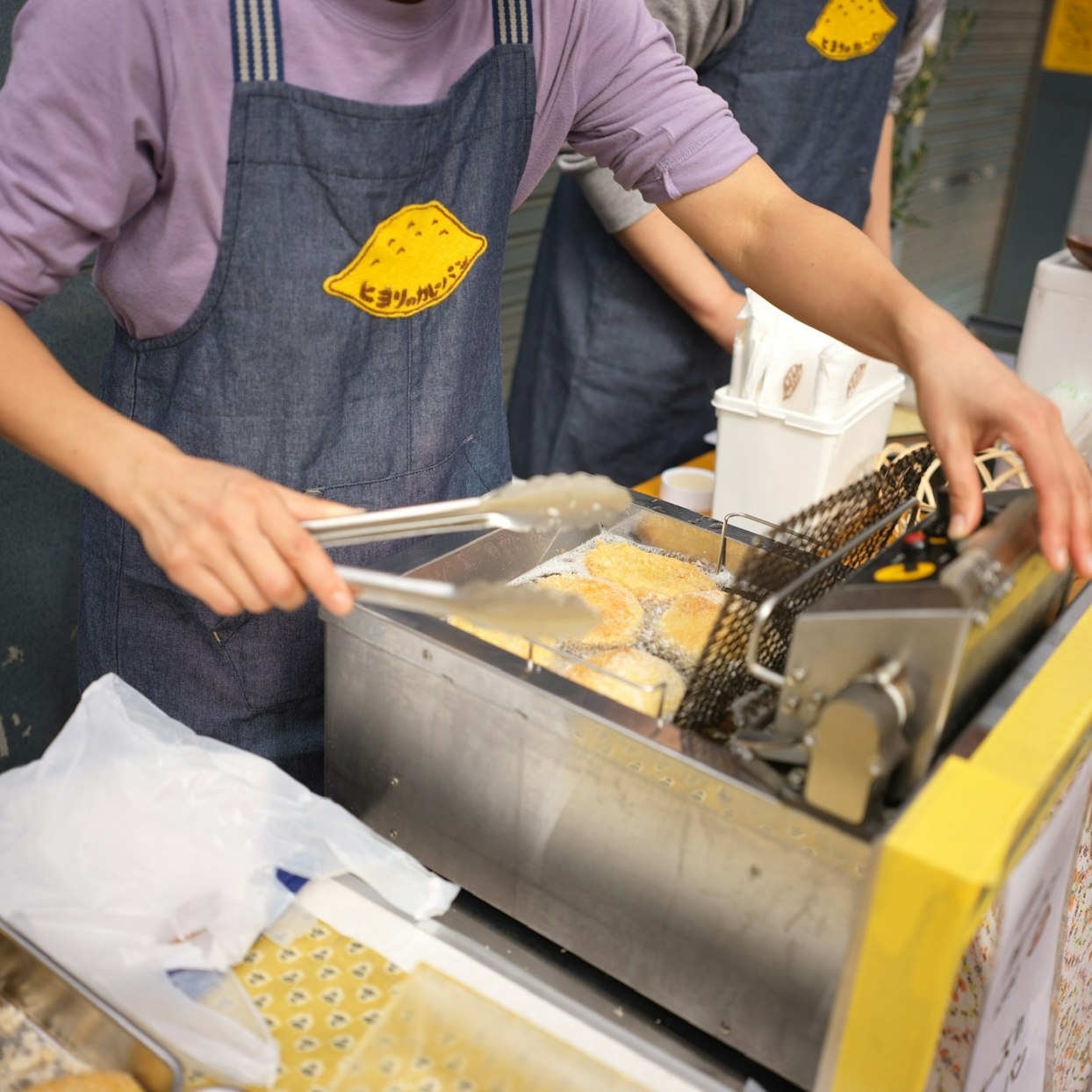 「ヒヨリノパン」のロゴが入ったデニムエプロンを着用した人物が、調理器具を使って揚げ物用のフライヤーでカレーパンを調理している。