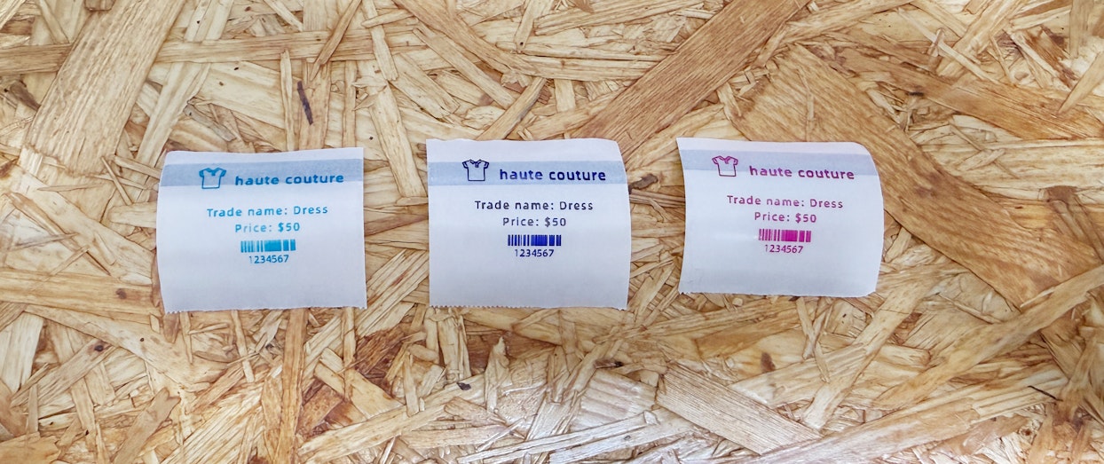 OSB合板の上に並べられた3枚の値札ラベル。各ラベルにはTシャツのアイコン、"haute couture"のロゴ、商品名（Trade name: Dress）、価格（Price: $50）、バーコードと番号（1234567）が印字されており、左から青緑色、紺色、マゼンタ色で色分けされている。