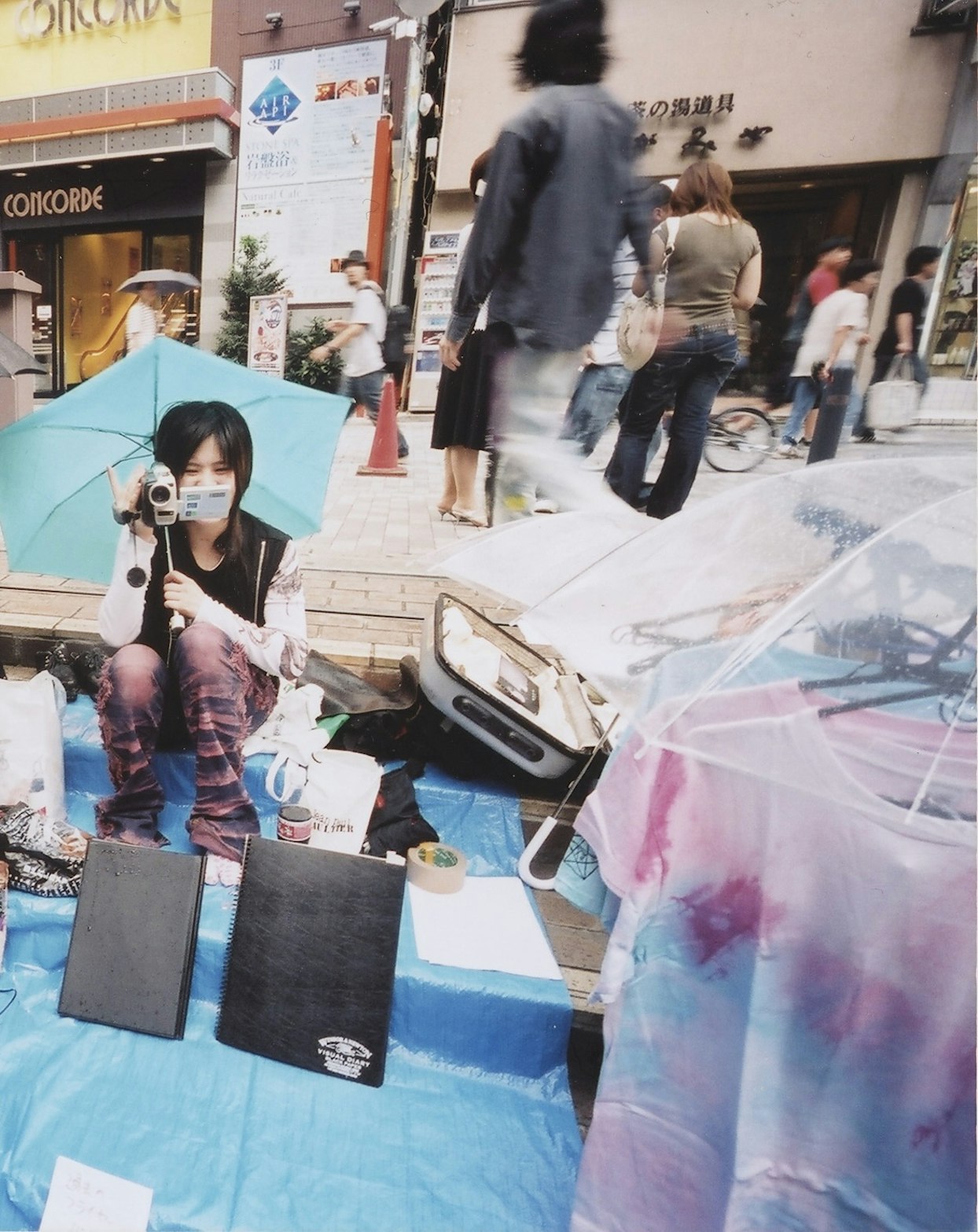 日本の街頭で、青い傘を持ちビデオカメラを構える若い人物。周りには通行人や商店が見える。地面には青いシートが敷かれ、その上に本やTシャツなどの商品が並べられている