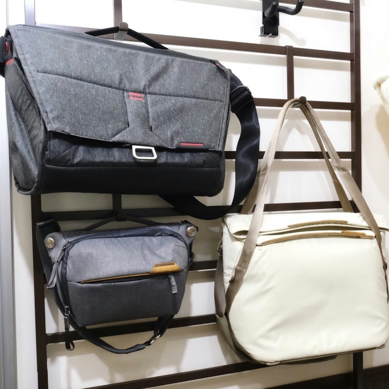 木製の棚に掛けられた3つの異なるバッグ。上部に大きな灰色のメッセンジャーバッグ、中央に小さな青灰色のカメラバッグ、下部に白いショルダーバッグが並んでいる。