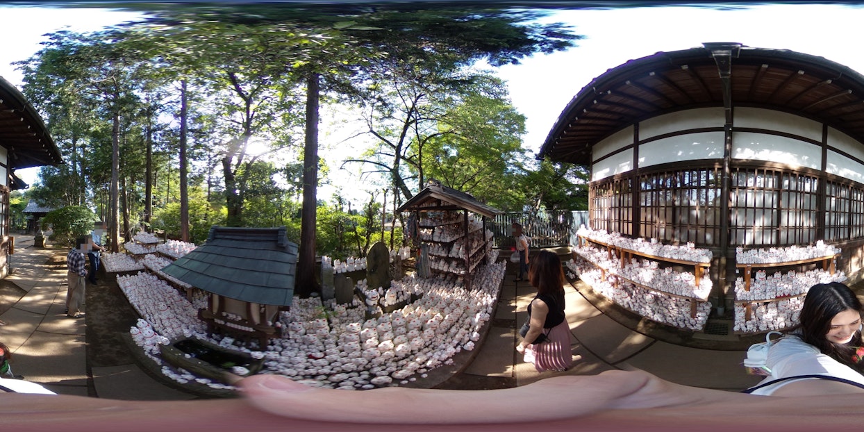 神社の境内で、絵馬や招き猫が大量に祀られた様子を360度パノラマで捉えた画像。木々に囲まれ、建物の一部や参拝客も見える。