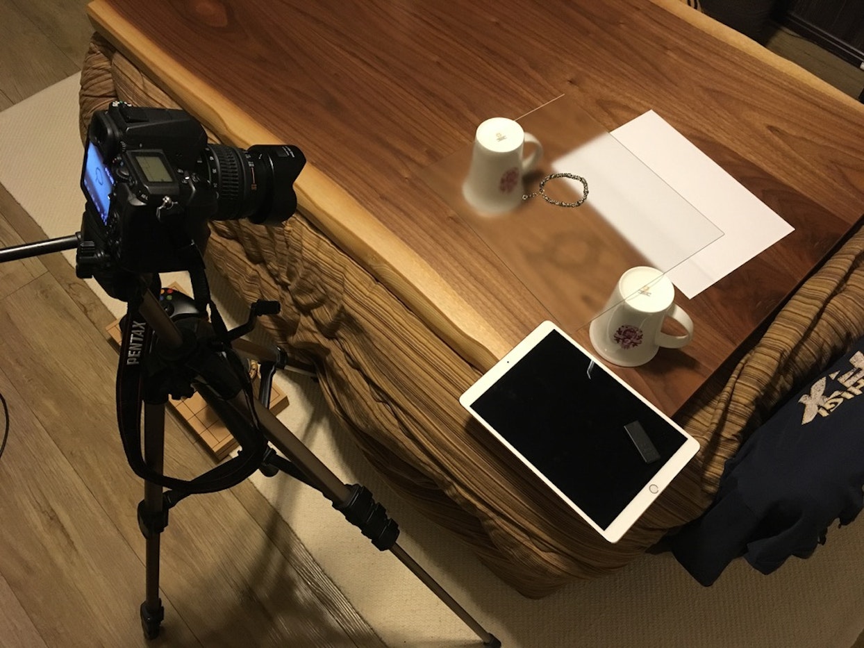 カメラ三脚、木製テーブル、2つのコーヒーカップ、タブレット、紙が置かれた写真撮影セットアップの様子