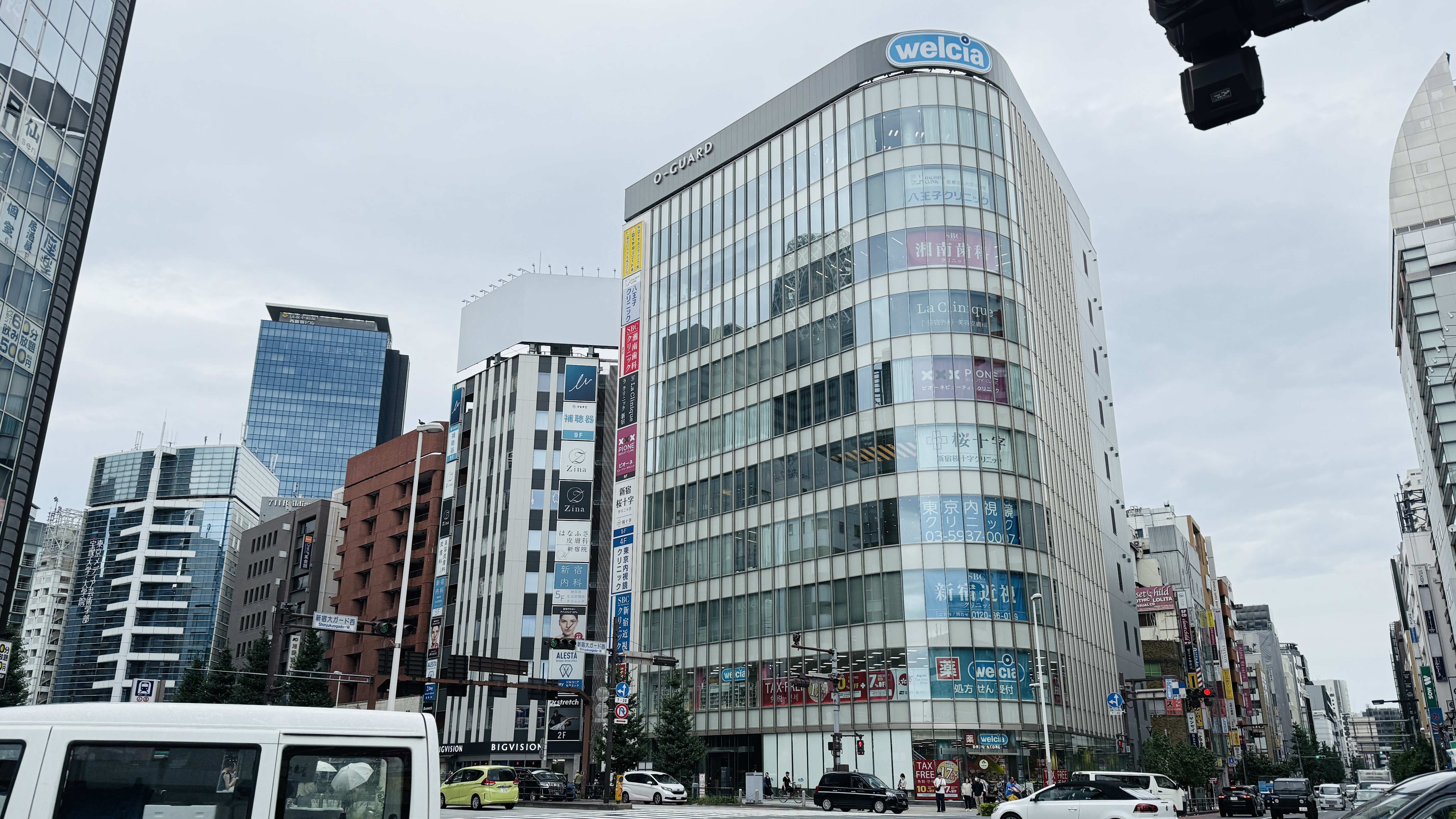 都心の交差点に建つ、ガラス張りの大きなビル。ビルの上部には「welcia」のロゴがあり、窓には様々な商品の広告が貼られている。手前には白いワンボックスカーや他の車が写っている。