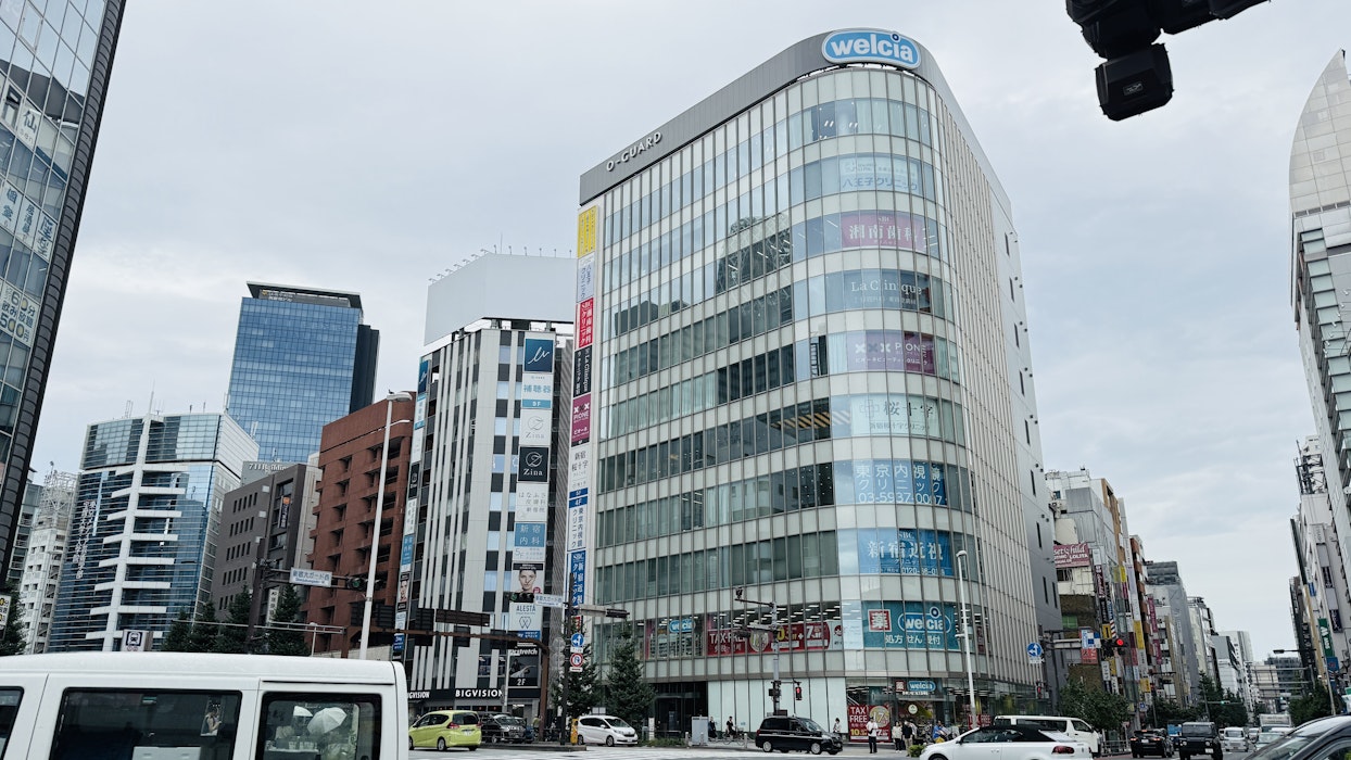都心の交差点に建つ、ガラス張りの大きなビル。ビルの上部には「welcia」のロゴがあり、窓には様々な商品の広告が貼られている。手前には白いワンボックスカーや他の車が写っている。