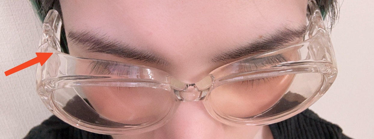  目元を拡大した画像で、一人の人物が透明な保護メガネを着用している。眉毛やまつ毛、額の一部が見え、左側のテンプルを指し示す赤い矢印が描かれている。