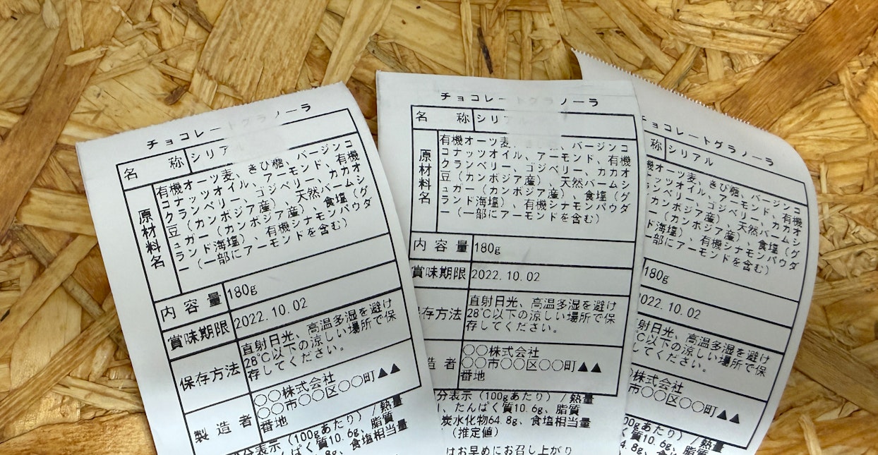 OSB合板の背景に並べられた、チョコレートグラノーラの食品表示ラベル3枚。3枚のうち2枚の印字が少し欠けている。