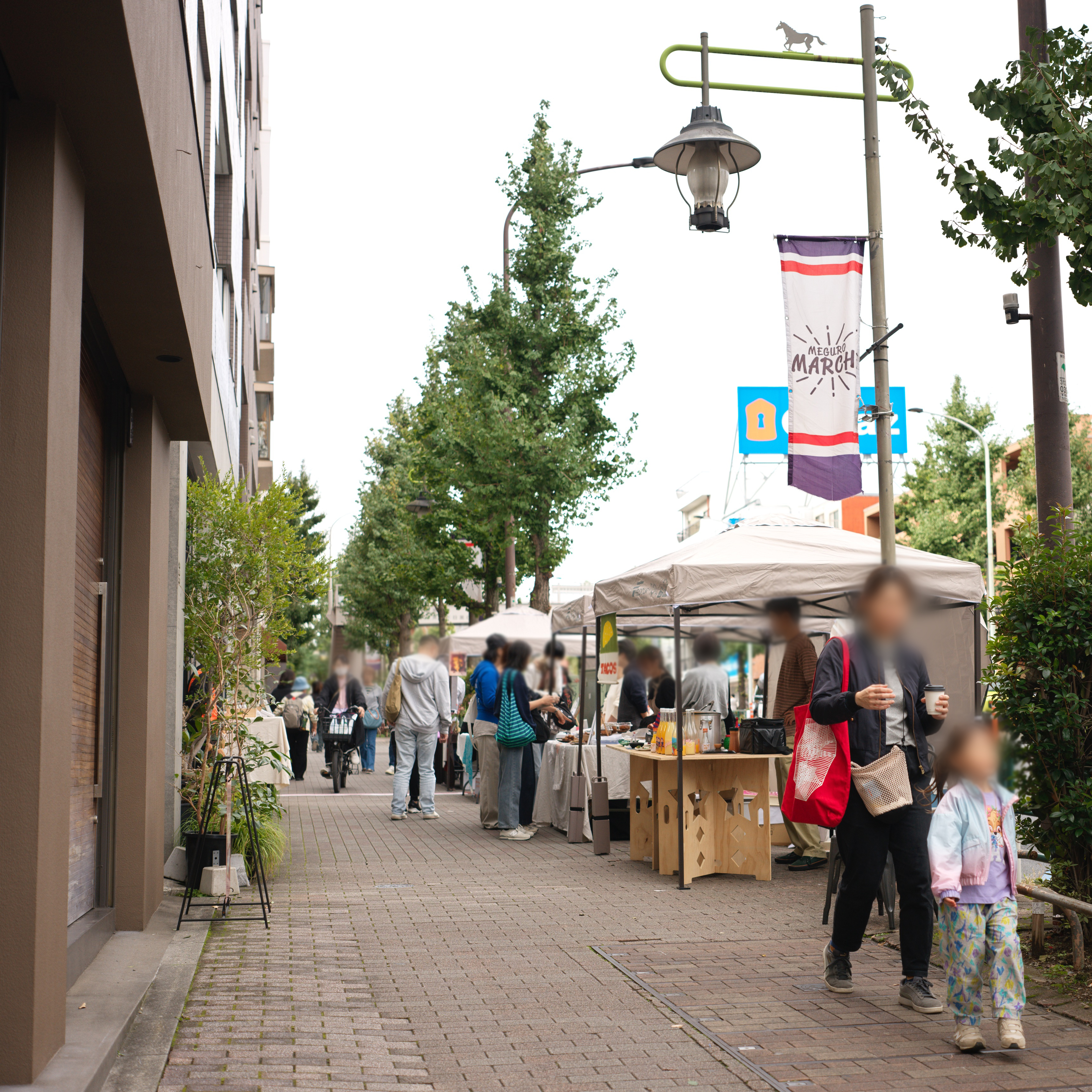 歩道で行われているマルシェの様子を捉えた写真。レンガ敷きの歩道沿いにテントがいくつか並び、多くの人が集まっている。右奥の街灯には「MEGURO MARCHE」と書かれたバナーが吊るされている。