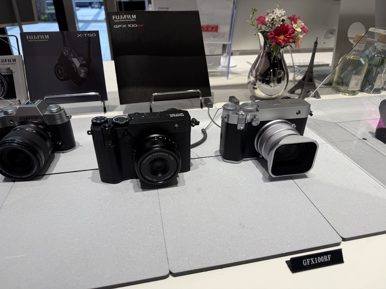 FUJIFILMのカメラ展示。X-T50とGFX100RFの製品カタログの前に、3台のデジタルカメラが並んでいる。背景には花瓶と小さなエッフェル塔のオブジェが見える。