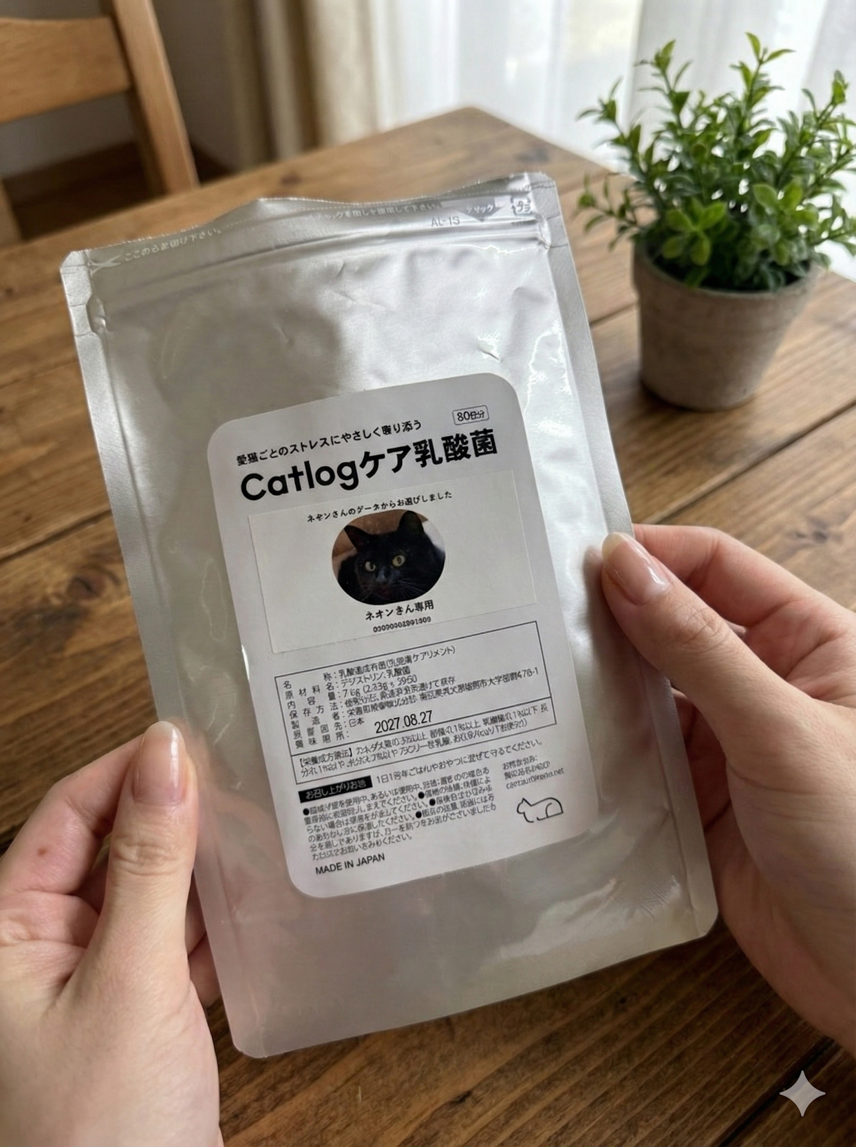 木目調のテーブルの上で両手に持たれた「Catlogケア乳酸菌」という猫用サプリメントの銀色のパッケージ。パッケージ中央のラベルには「愛猫ごとのストレスにやさしく寄り添う」というキャッチコピーに加え、黒猫の顔写真、「ネオンさん専用」という文字、「80日分」の表記、原材料名や賞味期限2027年8月27日などの詳細情報が記載されており、背景の右奥には小さな鉢植えの観葉植物が置かれている。