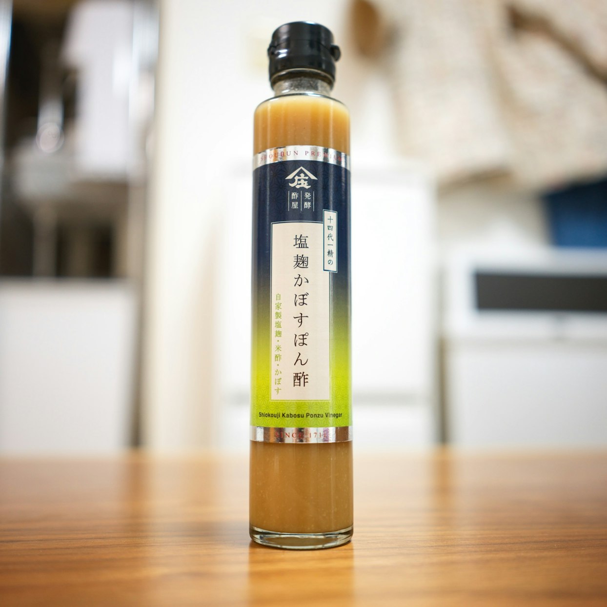 木製のテーブルの上に置かれたガラス瓶の調味料。「庄」のロゴと「十四代一精の塩麹かぼすぽん酢」という文字が縦にデザインされたラベルが貼られている。