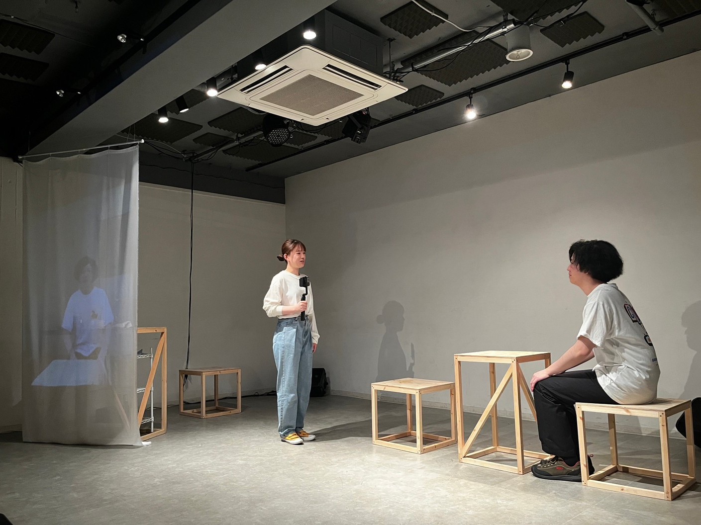 三鷹・SCOOLでの「シーユレーター」上演写真。出演者のかすがいこと子が、手持ちカメラを持ちながら森悟に話しかけている。左端のスクリーンには、舞台をカメラで写した映像が投影されている。