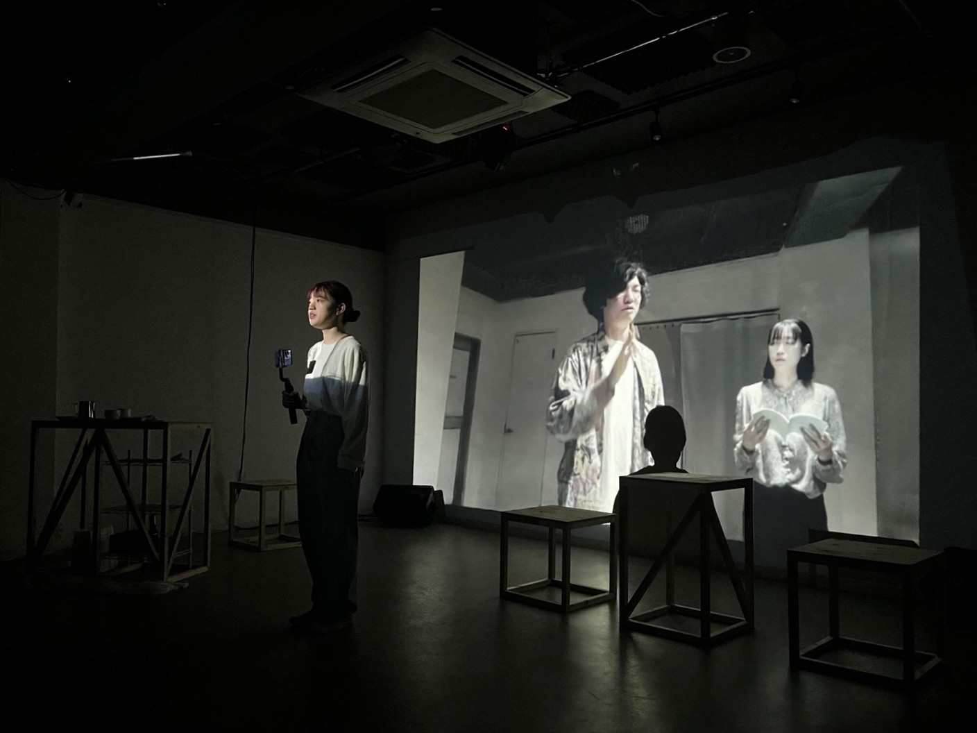 三鷹・SCOOLでの「シーユレーター」上演写真。暗い舞台中央にて、出演者のかすがいこと子が手持ちカメラを持ちながら正面を向いている。後方のスクリーンには、過去のシーンの映像が早回しで映し出されており、走馬灯のようにも見える。