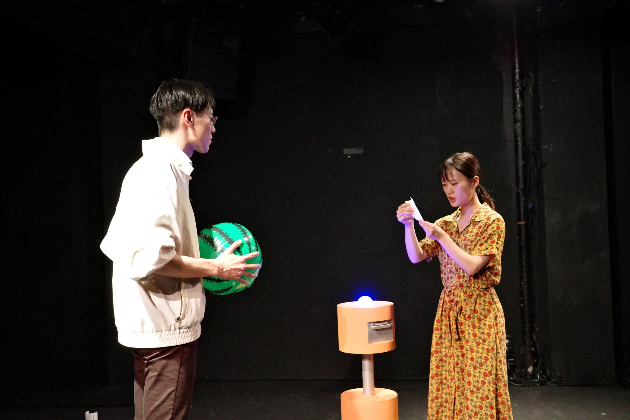 吉報 vol.1での「【炎上覚悟】海の家の裏側全部教えます」上演写真。出演者の高木彩加が感熱紙に印刷された文字を読む様子を、スイカ柄のビーチボールを手に持った山﨑陶熙が見つめている。二人の間には、子どもの背丈ほどのオレンジ色のロボットがたたずんでいる。