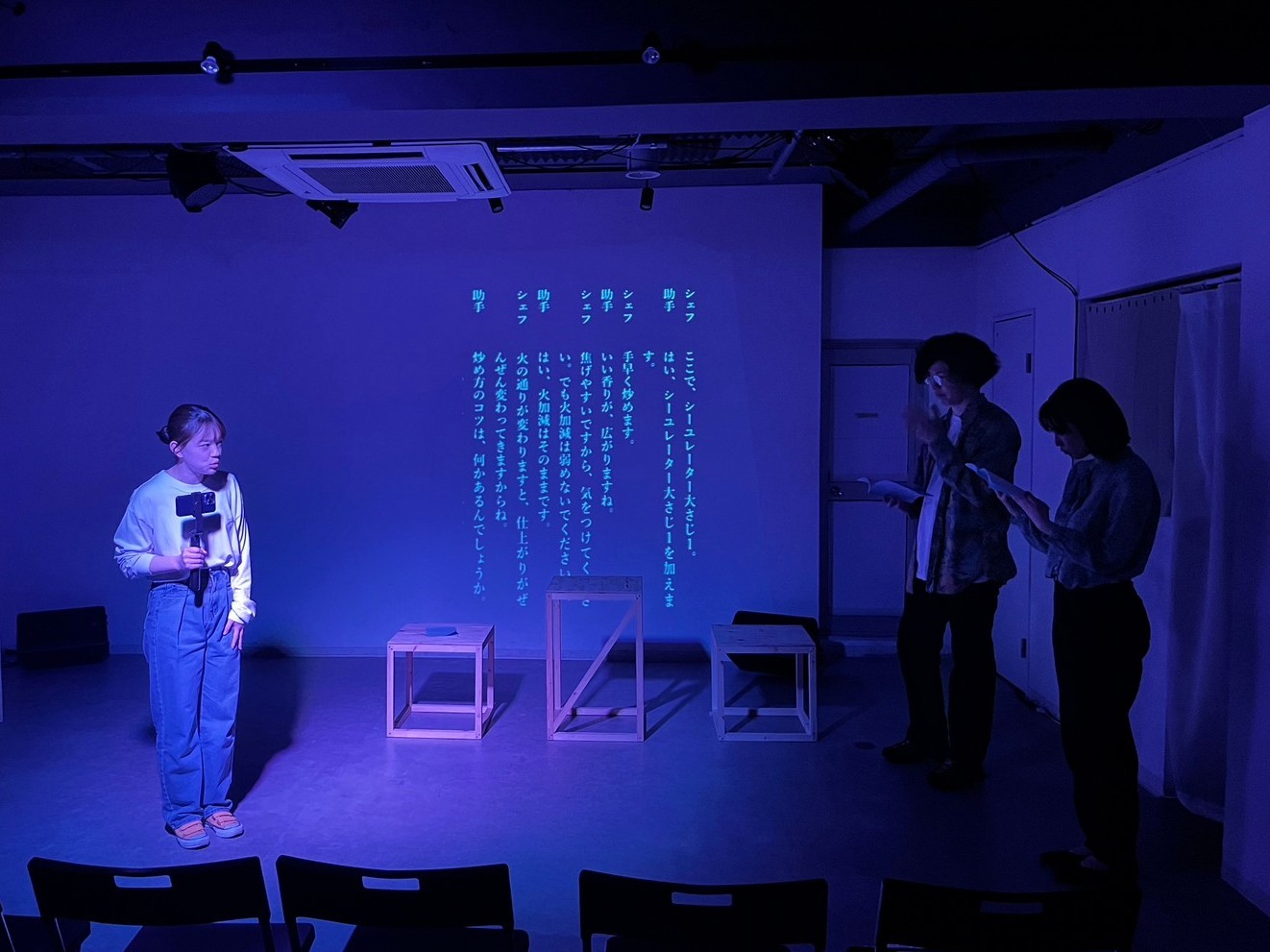 三鷹・SCOOLでの「シーユレーター」上演写真。青紫色に照らされた舞台中央にて、出演者のかすがいこと子が手持ちカメラを持ちながら劇中劇を披露しており、その様子を森悟と七星束子が舞台右端で確認している。後方のスクリーンには、台本のテキストが投影されている。