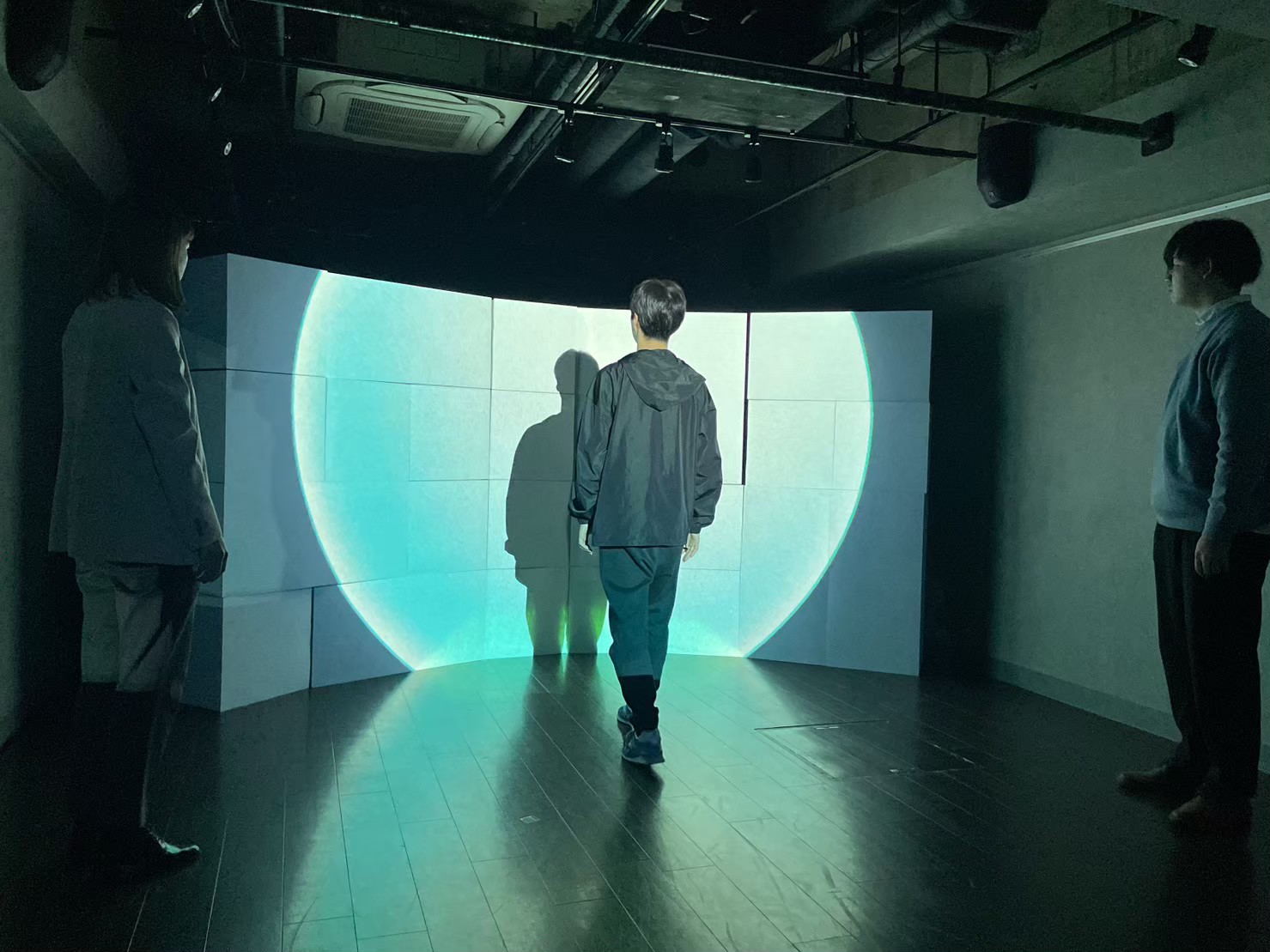 新宿眼科画廊での「球（kyu）」上演写真。舞台奥にて扇形に立てられた白い壁に、大きな球体が投影されている。舞台中央に出演者の佐藤昼寝、その両脇に新山志保と森悟が、それぞれ球体を見つめている。映像には、佐藤昼寝の影が意図的に大きく映りこんでいる。