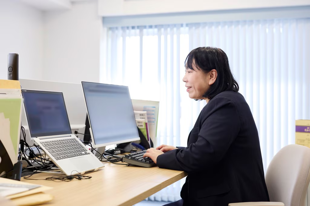 オフィスで、紺色のスーツを着た中年女性が、自身のデスクに座ってパソコン操作をしている様子。女性は笑顔を浮かべており、何かを確認しながらタイピングをしているように見えます。デスク上には、ノートパソコン、外部モニター、書類などが配置されており、窓際には垂直なブラインドを通して明るい光が差し込んでいます。