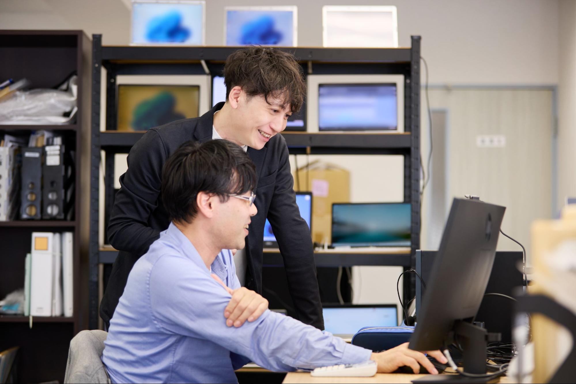 笑顔で肩に手を置くスーツ姿の男性が、デスクに座りモニターを見つめる眼鏡をかけた男性に、親身になって仕事の内容を説明したり、アドバイスを送ったりしている様子。オフィスの一角で、和やかな雰囲気の中、二人のチームメンバーが協力して業務に取り組んでいる様子が伝わります。背景には、いくつかのモニターと棚があり、活気ある職場環境がうかがえます。