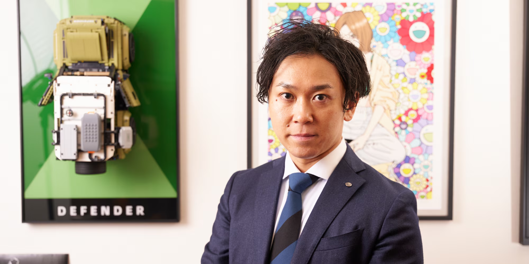 税理士の男性がオフィスで撮影された写真です。黒髪で、青いネクタイとネイビーのスーツを着用しています。背景には、2つの額縁が飾られています。左側にはレゴブロックでできたランドローバー・ディフェンダーの模型が、右側には村上隆の作品が飾られています。