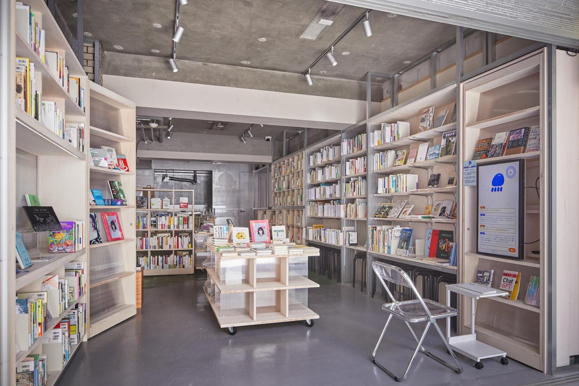 
書店内部を撮影した画像です。店内には床から天井まで届く木製の大きな本棚が並び、多種多様な本がずらりと並んでいます。手前には透明なパイプ椅子と、本を置ける小さなキャスター付きの台があります。その台にはタブレット端末が置かれ、青いキャラクターの画像と「チャットボット」の文字が表示されています。中央にはキャスターのついた低い平台があり、その上にも本が積まれています。天井は打ちっぱなしのコンクリートで、スポットライトが店内を照らしています。全体的に、明るくすっきりとしたモダンな内装の書店の様子がうかがえます。