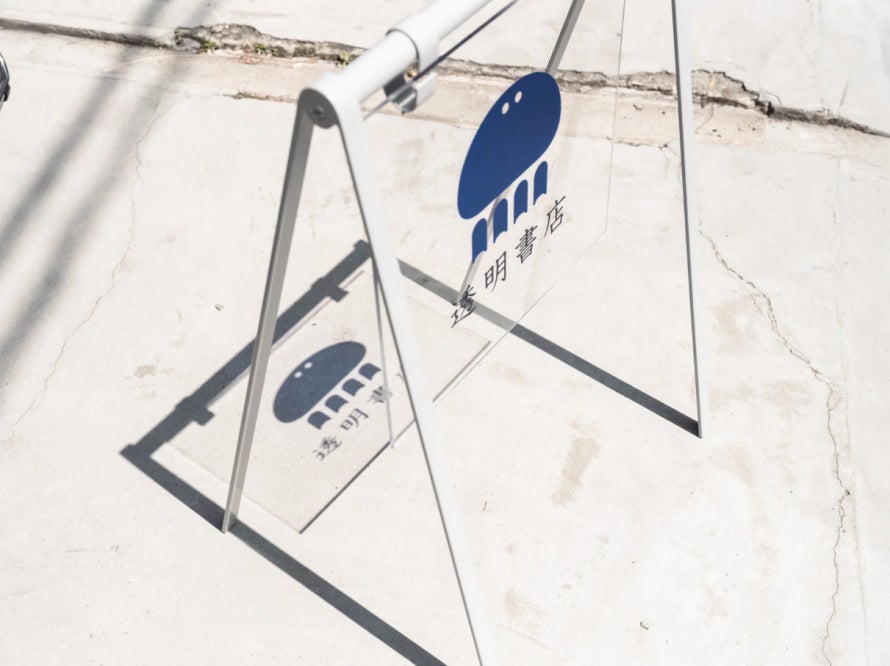 透明書店の立て看板。透明な板に青色のくらげのロゴと店名が記されており、地面に影が落ちている。