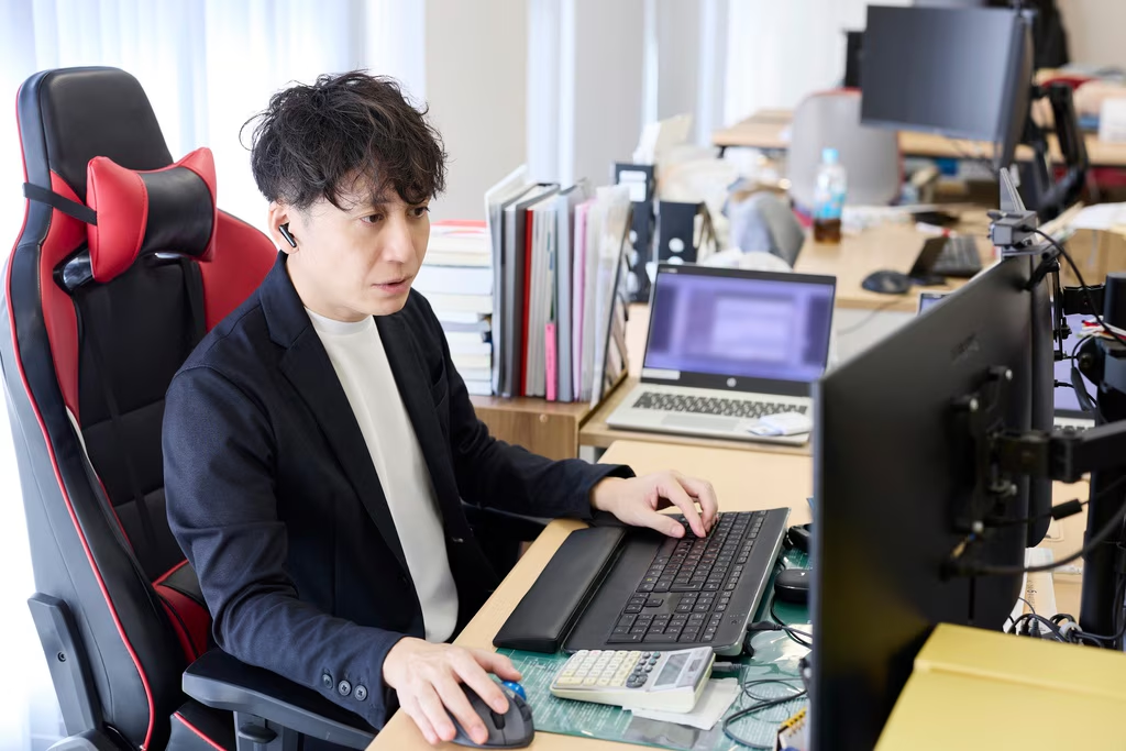 黒のジャケットを羽織り、ワイヤレスイヤホンを装着した男性が、赤と黒のゲーミングチェアのような椅子に座り、真剣な表情でデスクのモニターを見つめています。男性は両手をキーボードとマウスに置き、集中してPC操作を行っており、机上には電卓や書類、奥にはノートPCも見えます。背景には他のデスクやオフィス機器がぼかして写っており、オープンなオフィス環境で仕事に打ち込むビジネスパーソンの姿を表しています。