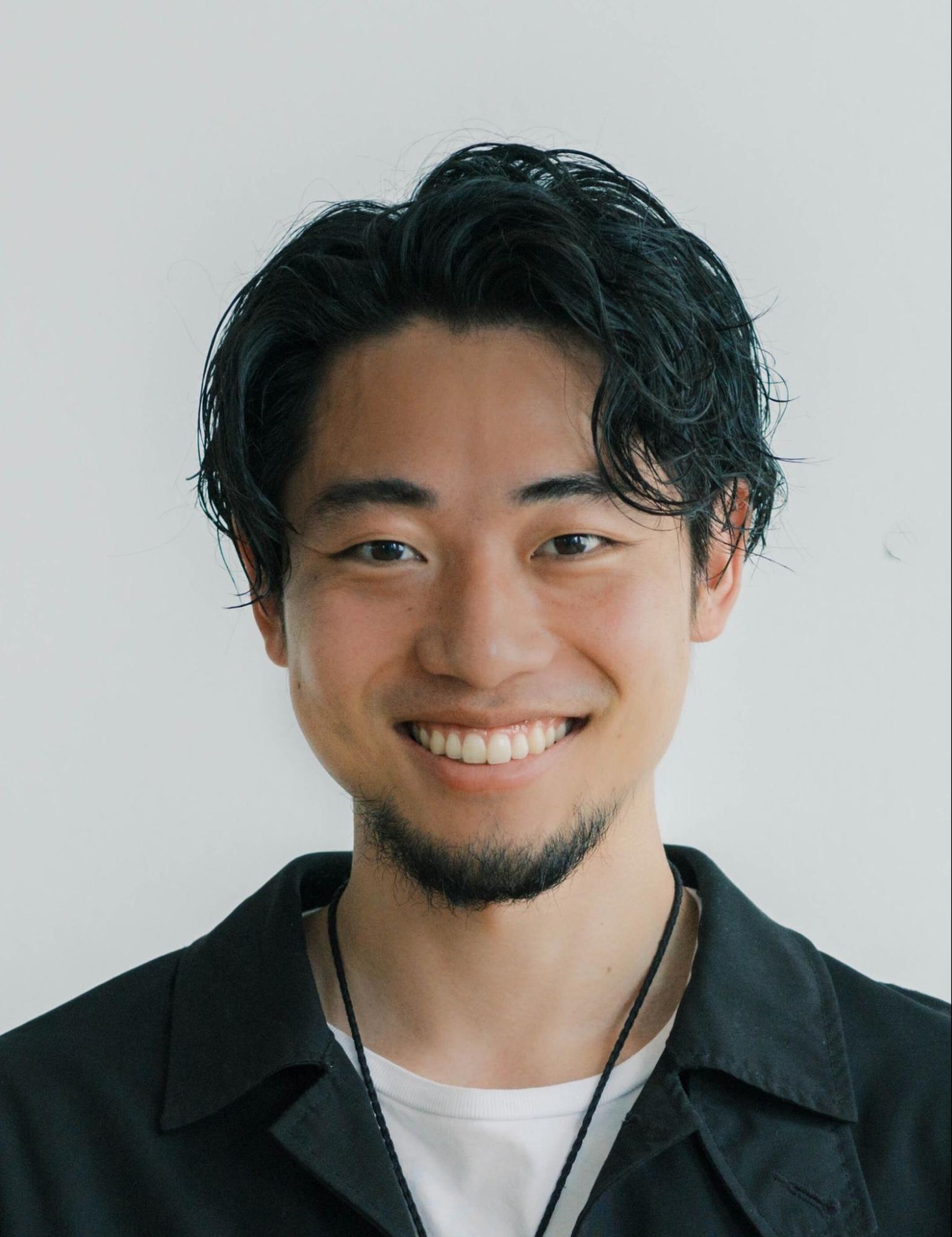 明るい白い背景の前で、黒い襟付きの服を羽織り、インナーに白いTシャツを着た若い男性が、カメラに向かって爽やかな笑顔を見せているバストアップ写真。男性はウェーブのかかった黒髪を自然に流し、顎には整えられた髭を蓄えており、首からは黒い紐のようなものを下げています。親しみやすさと誠実さを感じさせる表情が印象的なポートレートです。