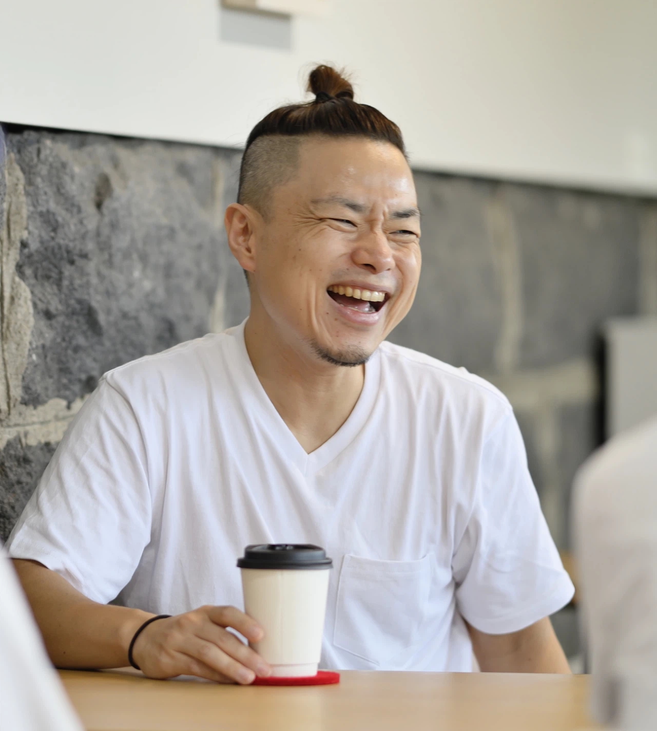 白いTシャツを着た男性が、笑顔で大きく口を開けて笑っている画像です。髪の毛は頭頂部で髷のように結んであり、刈り上げになっています。男性はコーヒーカップのような白い紙コップを手に持っており、テーブル越しに誰かと話している様子です。背景には石の壁が見えます