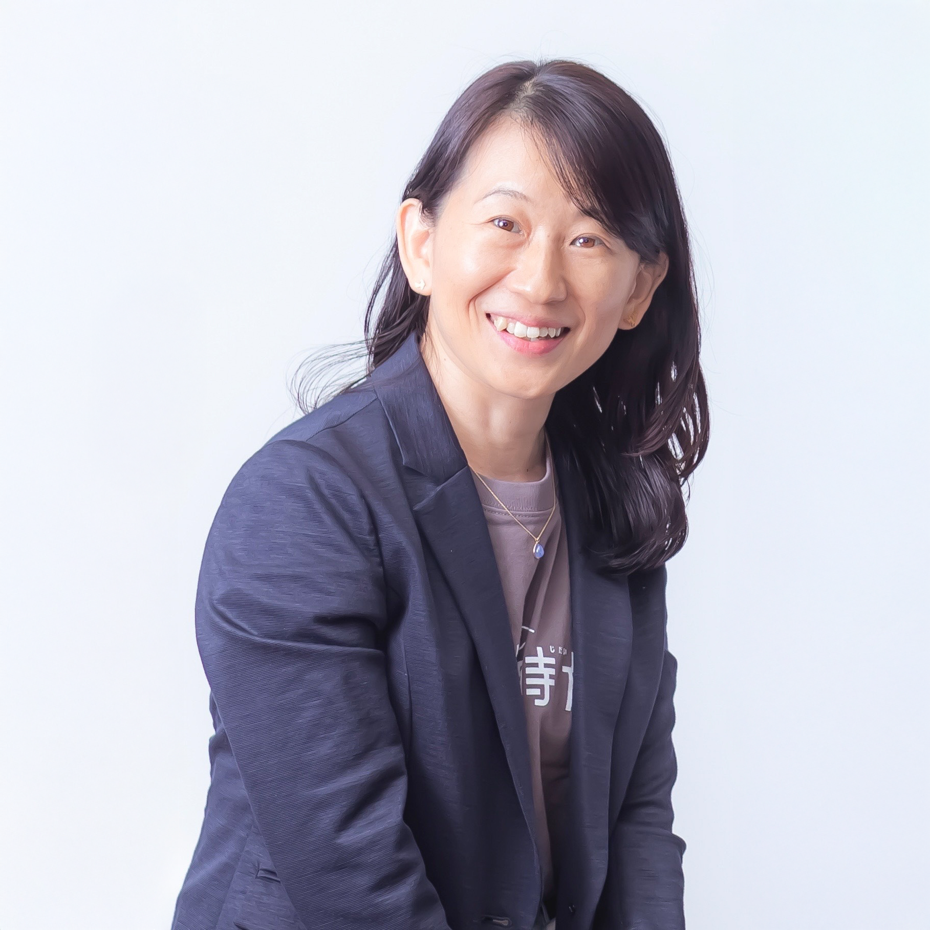 明るい白背景の前で、紺色のジャケットを羽織った女性が斜めに構えながら、穏やかで優しい笑顔をカメラに向けているポートレート写真。女性は暗い茶色のセミロングヘアで、ジャケットのインナーには文字の入った茶色のTシャツを着用し、小ぶりなペンダントが輝くネックレスを身に着けています。清潔感と親しみやすさが同居した、誠実な印象を与える一枚です。
