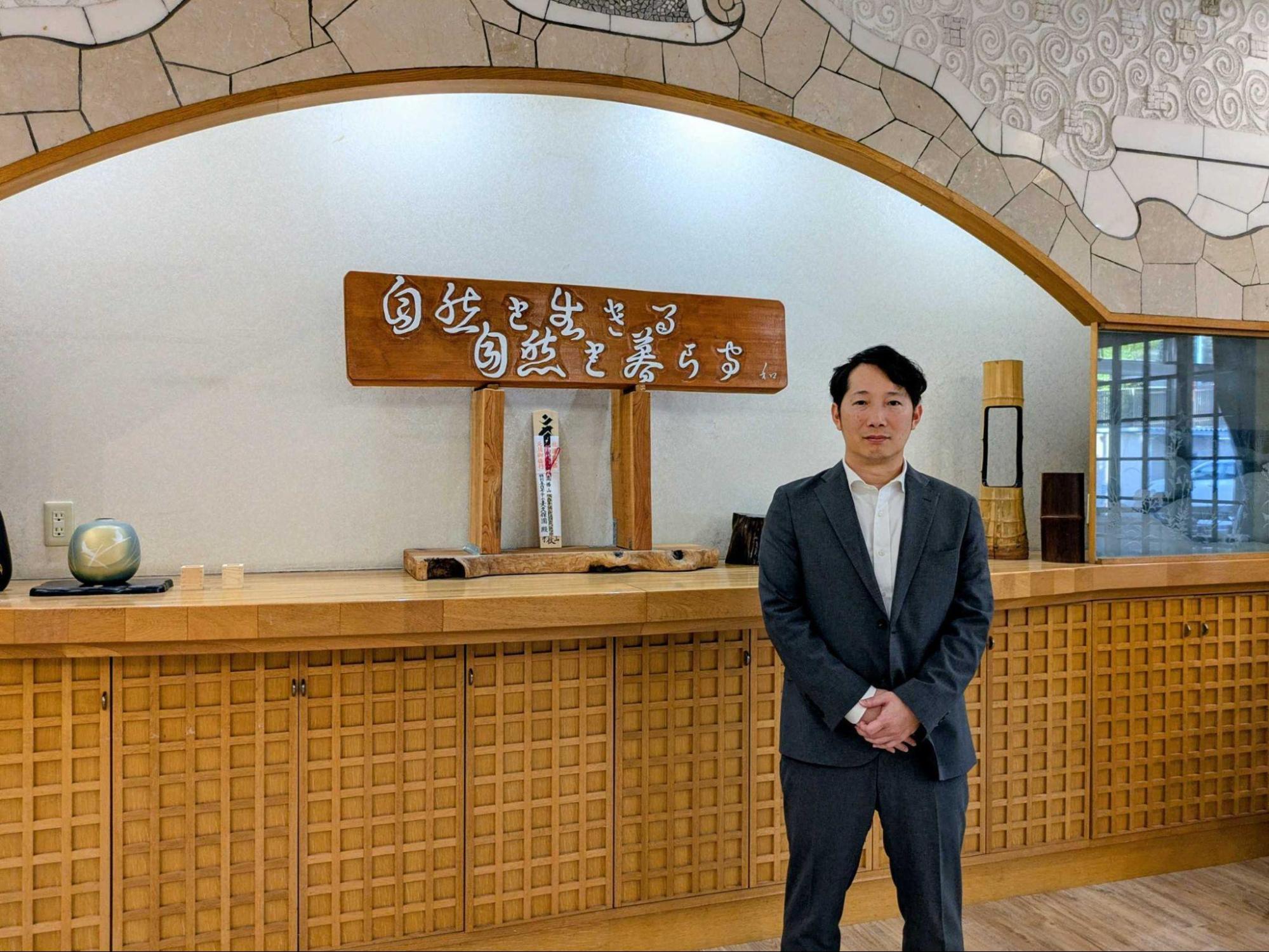 「自由を生きる 自然を暮らす」と書かれた木製の看板の前に立つ、グレーのスーツを着た男性の全身写真。背景には和風の格子戸の棚がある。