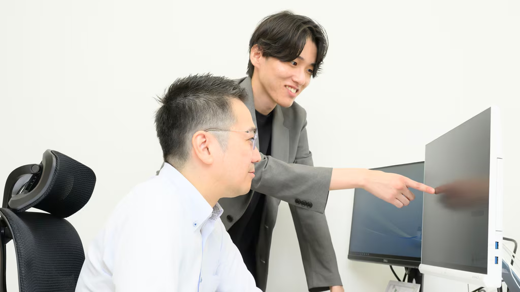 明るいオフィスのような場所で、白いシャツを着た男性がデスクの椅子に座り、その横でグレーのジャケットを羽織った若い男性が笑顔でパソコンの画面を指差しながら説明をしています。座っている男性は画面を真剣に見つめており、二人が仕事の打ち合わせや相談をスムーズに行っている様子が伝わってくる一枚です。