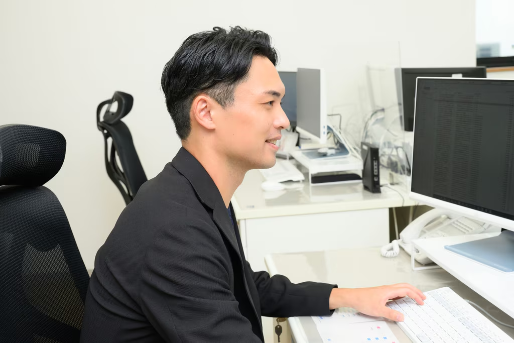 黒いレザーのオフィスチェアに座ったグレーのジャケット姿の若い男性が、正面を見据えながら熱心に話をしている様子です。左手を顔の横で大きく広げ、何かを説明するかのような力強いジェスチャーを交えており、インタビューや対談の中で自身の考えを生き生きと伝えている瞬間を捉えています。