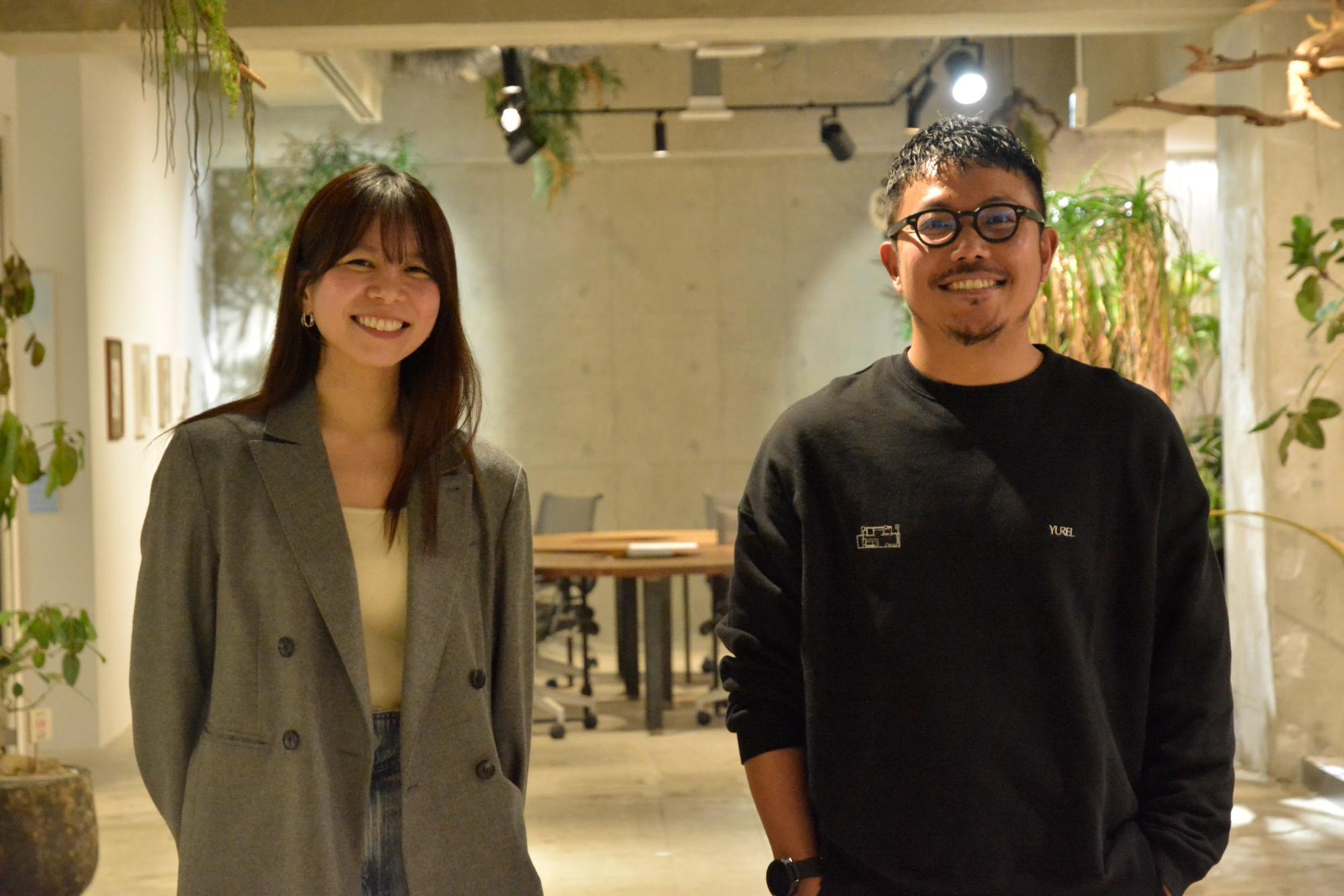コンクリート打ちっぱなしの壁や観葉植物がおしゃれなオフィス空間で、グレーのジャケットを着た笑顔の女性と、黒いスウェットを着て眼鏡をかけた笑顔の男性が並んで立っている写真です。背景には木製のテーブルやデスクチェアが見え、温かみのある照明が落ち着いたモダンな雰囲気を醸し出しています。