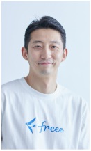 白いTシャツを着て微笑むfreee専務執行役員CHROの川西康之氏の写真です。Tシャツの胸元にはfreeeのロゴと文字がデザインされており、カジュアルな装いです。背景は白い壁で、左側に緑の観葉植物が写っています。