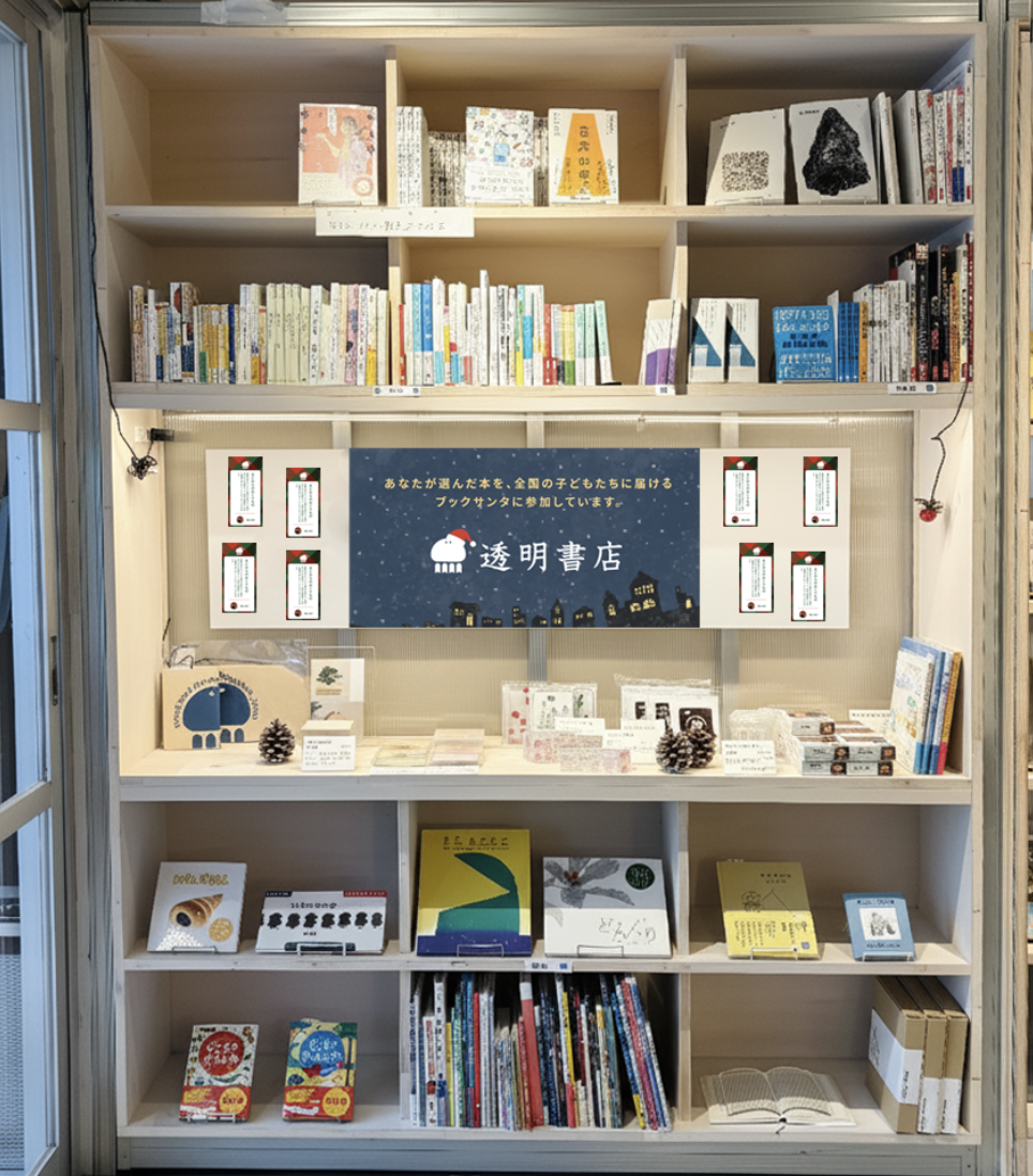 書店の木製の陳列棚を写した画像です。棚の中央には、夜空に家々がシルエットで描かれ、白いキャラクターと「透明書店」という文字が配置�されたポスターがあります。そのポスターの左右には、白い枠に赤と黒の縦線が入ったデザインのカードが複数並べられています。ポスターの上には、「あなたが選んだ本を、全国の子どもたちに届けるブックサンタに参加しています。」というメッセージが書かれています。棚には、本やカード、雑貨などがきれいに陳列されています。ポスターのすぐ下の段には、松ぼっくりや、透明のビニール袋に入ったギフトセットのような商品が見えます。さらにその下の段や一番下の段にも、様々な書籍や商品が並んでいます。棚の隅には、小さな蜘蛛の飾りもいくつか見られます。全体的に、温かみのある雰囲気の書店の様子が伝わってきます。