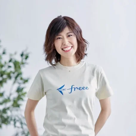 白っぽい背景の前で、胸にfreeeのロゴがプリントされた薄いクリーム色のTシャツを着た、笑顔の女性が写っています。女性はショートボブの髪型で、カメラに向かって優しく微笑みかけており、両手を体の後ろかポケットに入れているように見えます。左奥には、緑の植物の一部が少しだけ写り込んでいます。
