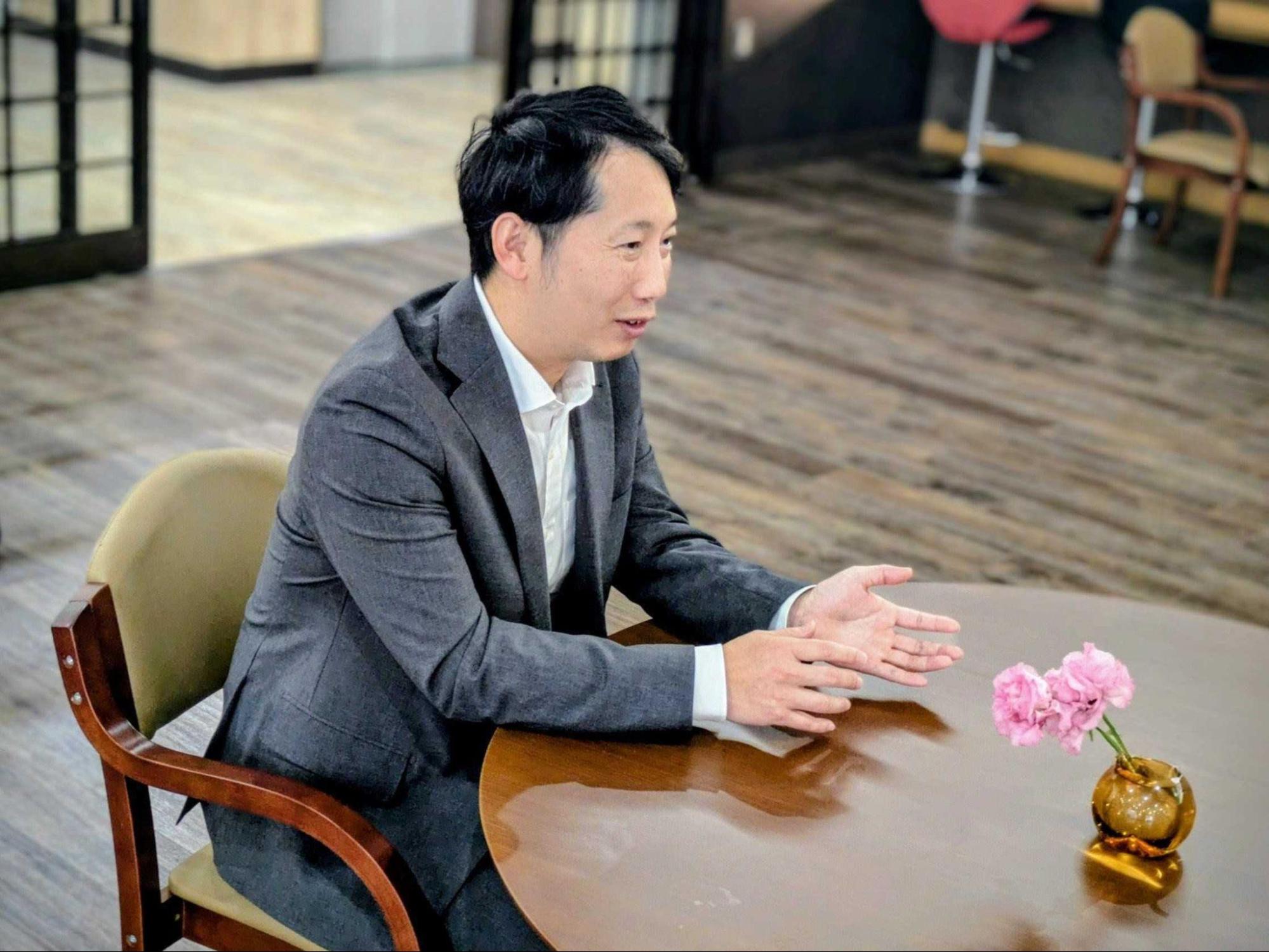 木製の 丸テーブルに座り、身振り手振りを交えながら横を向いて話すスーツ姿の男性。テーブルの上には小さなピンクの花が生けられた一輪挿しがある。