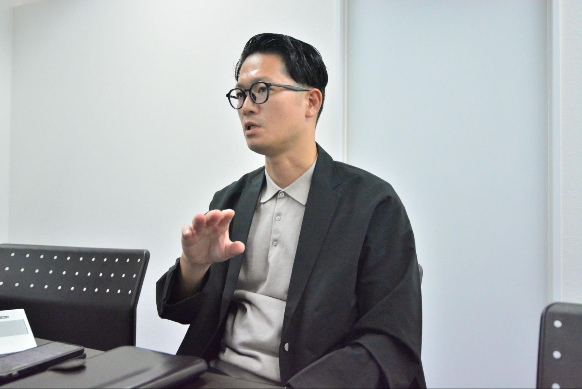 黒いジャケットに明るい色のポロシャツを着た眼鏡の男性が、白い壁を背景にした会議室で、身振り手振りを交えながら熱心に話している様子を捉えた上半身のクローズアップ写真です。手前にはテーブルの一部や黒い椅子、スマートフォンらしきものが写っています。