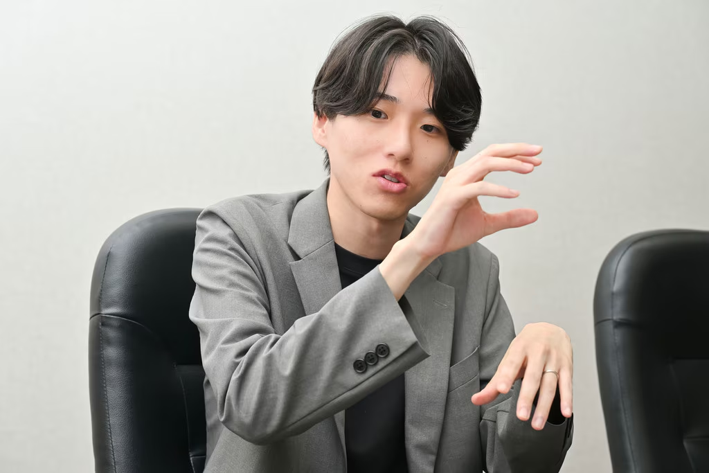 黒いオフィスチェアに座り、グレーのジャケットを身にまとった若い男性が、正面を向きながら身振り手振りで熱心に話をしている様子です。左手を胸の高さまで上げ、何かを掴むようなジェスチャーを交えながら、真剣かつ穏やかな表情で言葉を紡いでおり、インタビューや対談のワンシーンを切り取ったような躍動感のある一枚となっています。