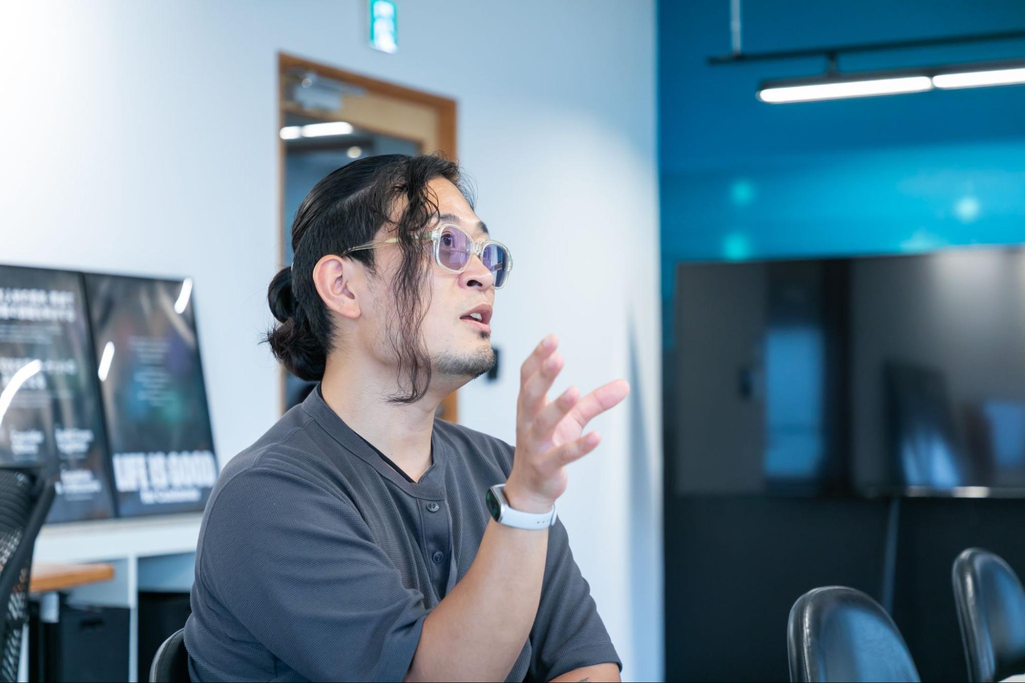 この写真は、会議室らしき場所で話している、サングラスをかけた長髪の男性を写しています。彼は無地のTシャツを着ており、何かを熱心に説明しているかのように、片手を上げて身振り手振りを交えています。背景には、大きなモニターやポスター、そしてシンプルな椅子がいくつか見えます。