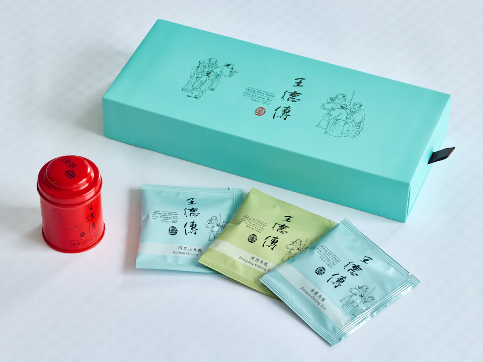 王德傳のお茶の写真。右上：王德傳のお茶の箱。水色の地の上、中央には王德傳のロゴ、その両脇には古風な人物画があしらわれている。全体的に余白を残したデザイン。右下：王德傳のお茶のパックが3つ並んでいる。左から阿里山烏龍、凍頂烏龍、金萓烏龍。箱を踏襲したようなデザイン。　左下：王德傳のお茶の缶。全体を赤で塗られており、側面には王德傳のロゴ、蓋には印鑑で押したようなデザインが施されている。