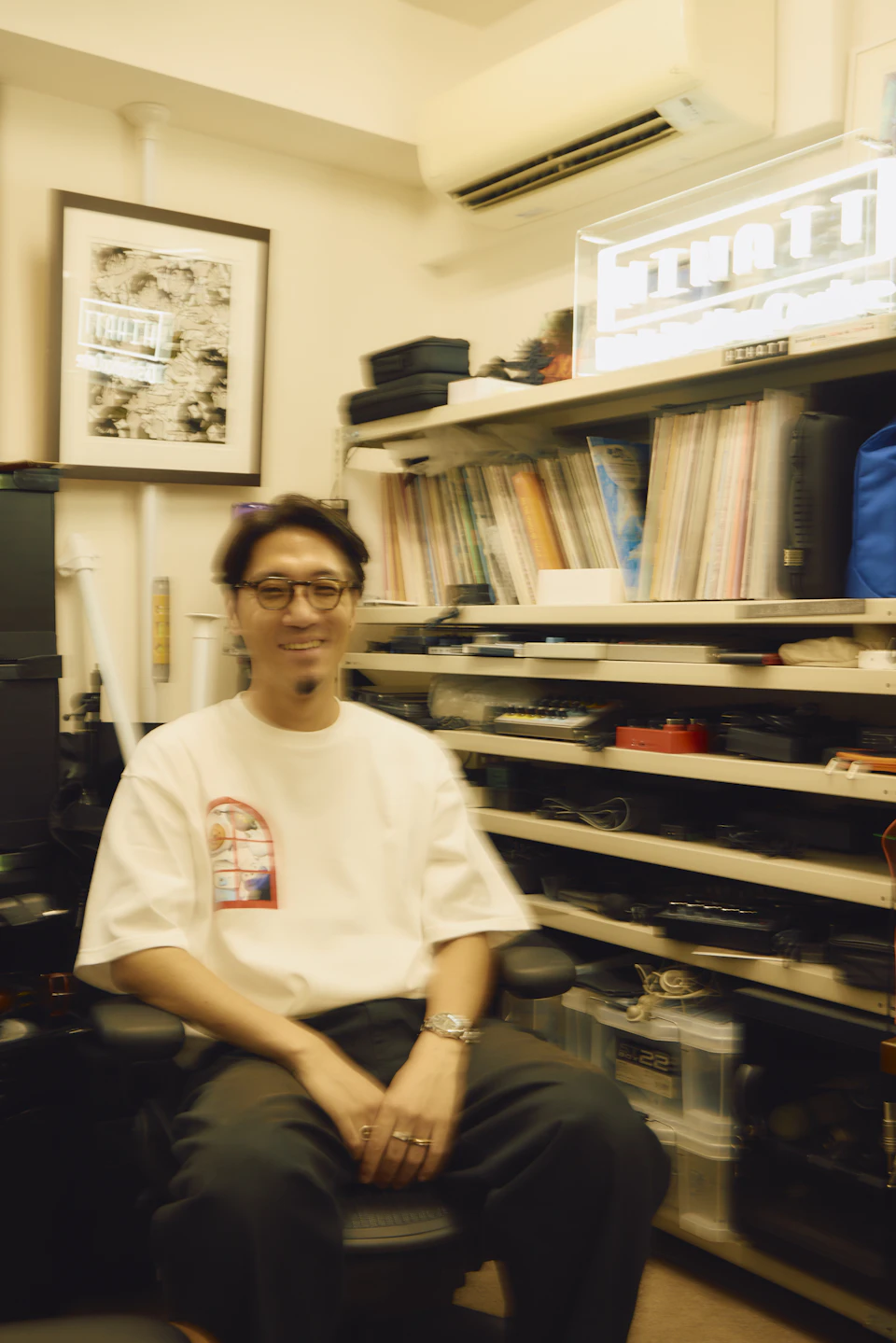 tofubeatsさんの作業部屋。数多くの作曲用の機器やファイルといった様々なものが収納されており、壁には縦50cm、横40cm程度の現代アートが額縁に入れて飾られている。部屋の中央でワーキングチェアに腰掛けながらtofubeatsさんが微笑んでいる。