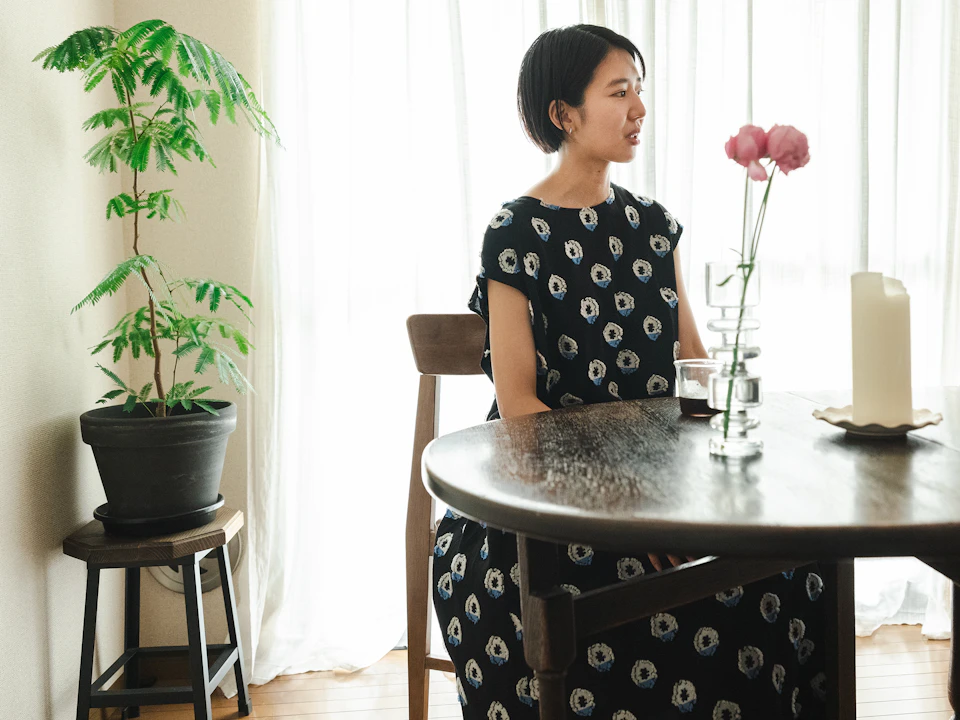 ご両親から受け継いだというダイニングテーブルと椅子の側で思い出を語る石山さん。テーブル、椅子ともに木製で、色はダークブラウン。テーブルの上にはコップと、花が二輪挿された花瓶と、太いキャンドルのようなものが置かれている。