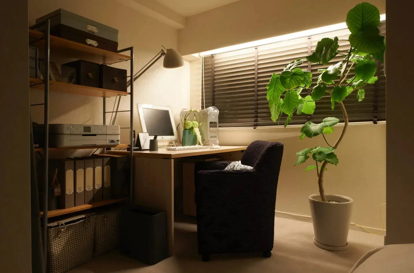 寝室の一角にある書斎コーナー。デスクと椅子、背の高い観葉植物が置かれ、窓には間接照明で照らされたウッドブラインドが掛かっている。