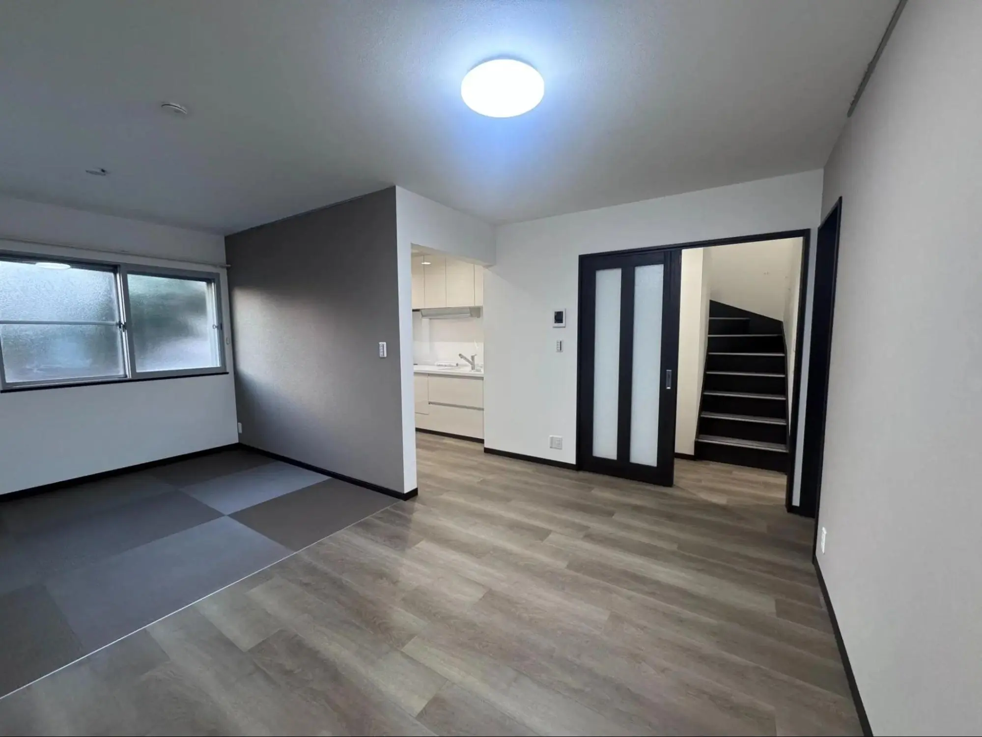 リノベーション後のLDK写真。床は木目調フロアタイルで、奥にキッチン、右手に階段へ続く黒枠の引き戸が見える。