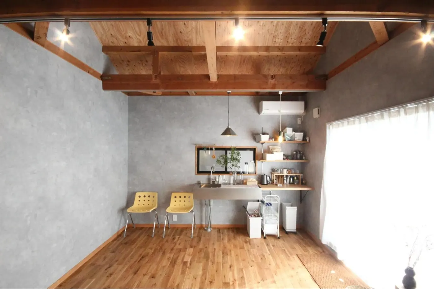 構造用合板と梁を現しにした高い天井のアトリエ。モルタル風のグレーの壁にステンレス製の壁付けシンクを設置し、床はオーク無垢材で仕上げている。黄色いプライウッドチェアが映える、素材の質感を活かした和洋折衷のリノベーション内観。アズ建設の事例。