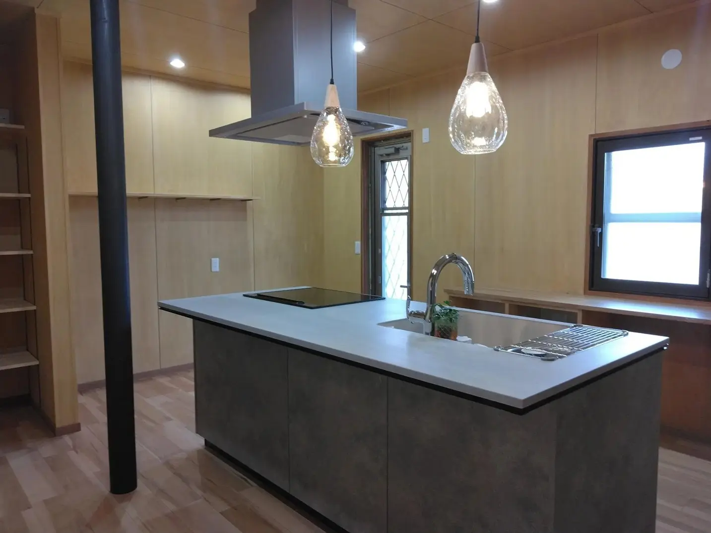 シナベニヤの天井と壁、無垢フローリングで仕上げられた、木の温もりあふれる対面キッチン。段差を解消したバリアフリー設計で、手前のダイニングから奥まで回遊できる動線が確保されている。奥の勝手口には断熱性能を高める内窓が設置されているのが見える。